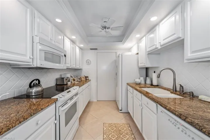 Kitchen with dark stone counters, white appliances, a raised ceiling, sink, and white cabinets
