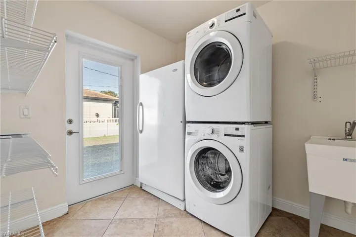 Inside Laundry room with light tile patterned flooring, sink, and stacked washer and dryer, freezer and plenty of shelving. Launry room is huge with exterior door leading to outside yard