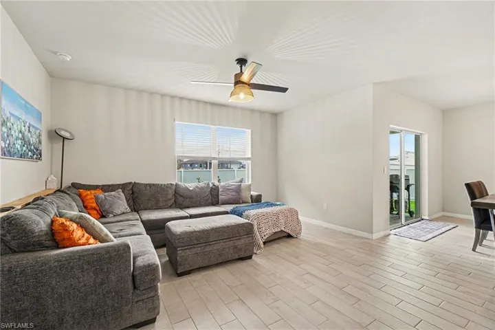 Spacious living area featuring light-toned walls, wood-finish flooring, a ceiling fan with integrated lighting, and a large window with horizontal blinds