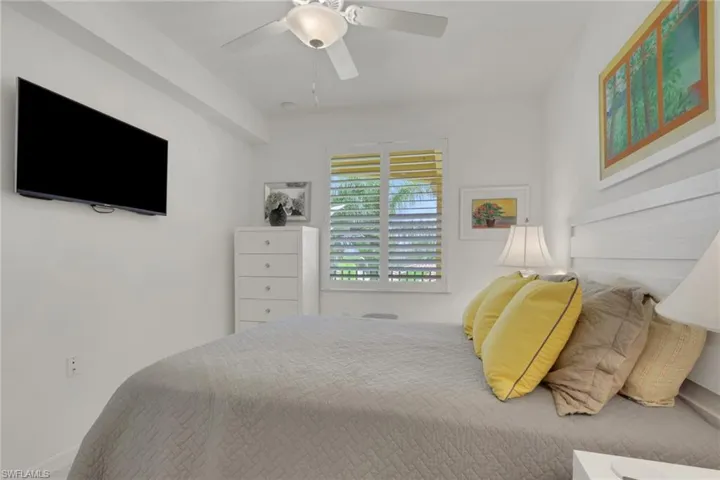 Bedroom featuring a wall-mounted television, ceiling fan, and window with plantation shutters