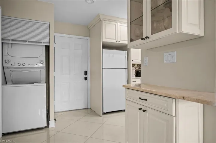 Kitchen with freestanding refrigerator, light tile patterned floors, glass insert cabinets, stacked washer and clothes dryer, and white cabinets