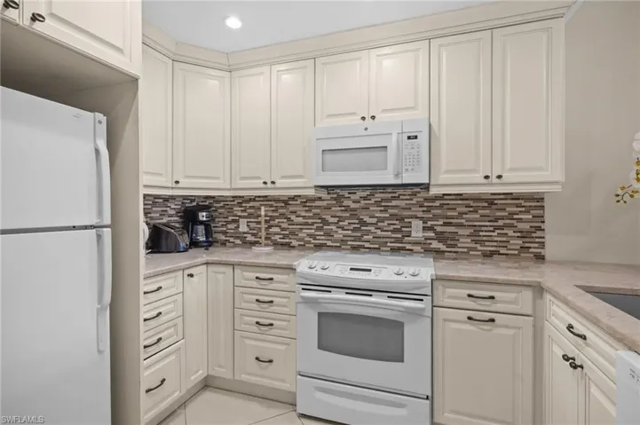 Kitchen featuring white appliances, light tile patterned flooring, tasteful backsplash, light stone counters, and white cabinets