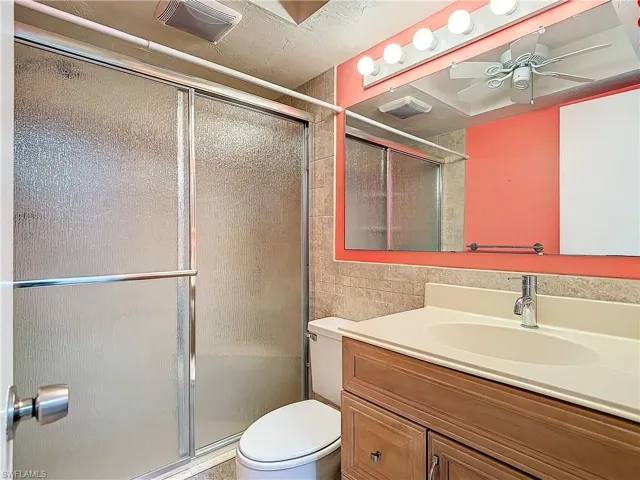 Bathroom featuring a shower with shower door, tile walls, toilet, vanity, and ceiling fan