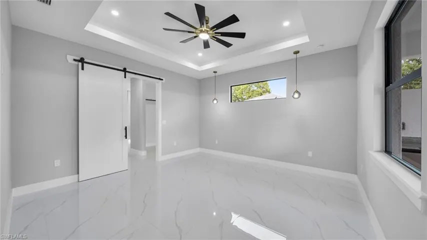 Empty room with a barn door, a raised ceiling, light marble finish flooring, recessed lighting, and ceiling fan