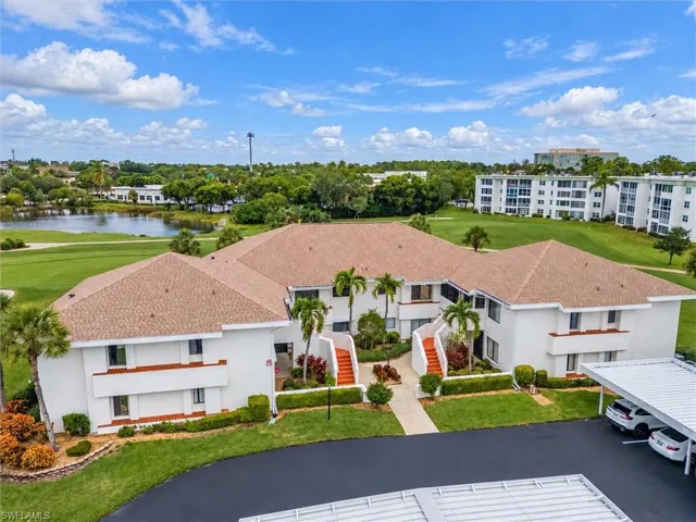 Multi-story building featuring a textured roof, white stucco exterior, exterior staircases, and attached carport parking, situated on grounds including manicured turf and a pond