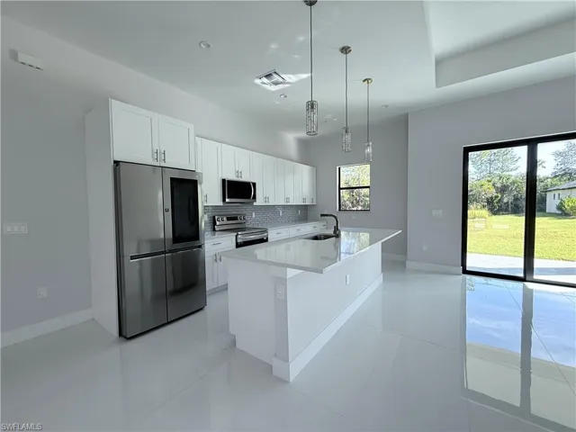 Kitchen featuring appliances with stainless steel finishes, white cabinetry, hanging light fixtures, a center island with sink, and tasteful backsplash