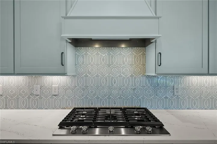Kitchen view of custom exhaust hood, stainless steel gas cooktop, light stone countertops, and tasteful backsplash