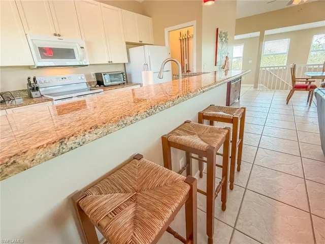 Kitchen with light tile patterned floors, light stone counters, white appliances, white cabinetry, and a kitchen breakfast bar