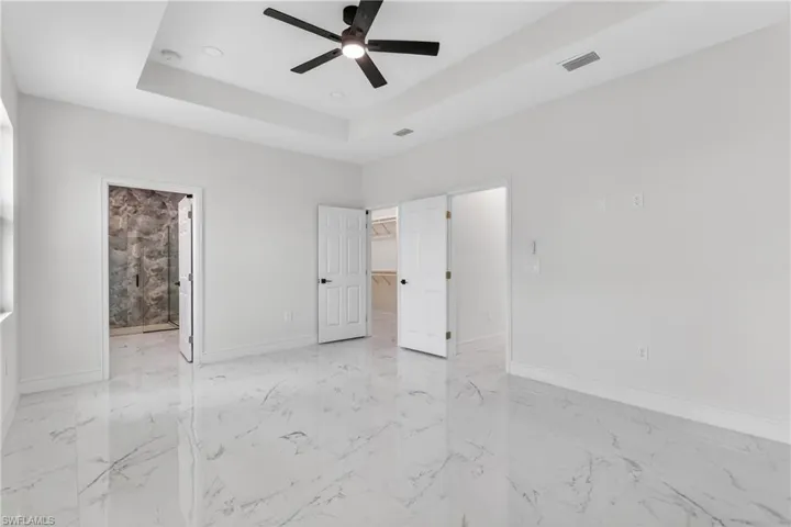 Unfurnished bedroom with a walk in closet, ensuite bathroom, a ceiling fan, a tray ceiling, and light marble finish floors