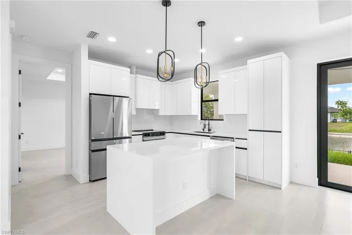Modern kitchen featuring a large central island with a waterfall edge, stainless steel refrigerator, contemporary pendant lighting, white high-gloss cabinetry, and a full-light glass door