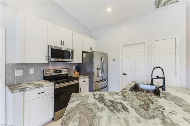 Kitchen featuring appliances with stainless steel finishes, light stone counters, white cabinetry, vaulted ceiling, and backsplash