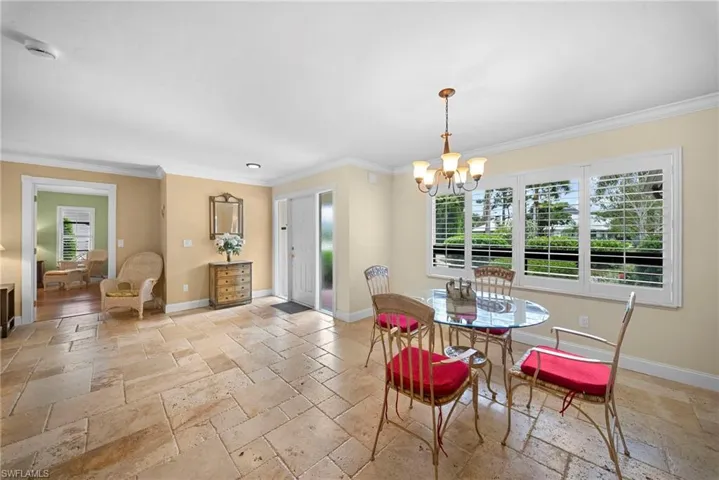 Textured stone flooring, crown molding, white paneled entry door with sidelight, large window with white plantation shutters, and a chandelier with glass shades