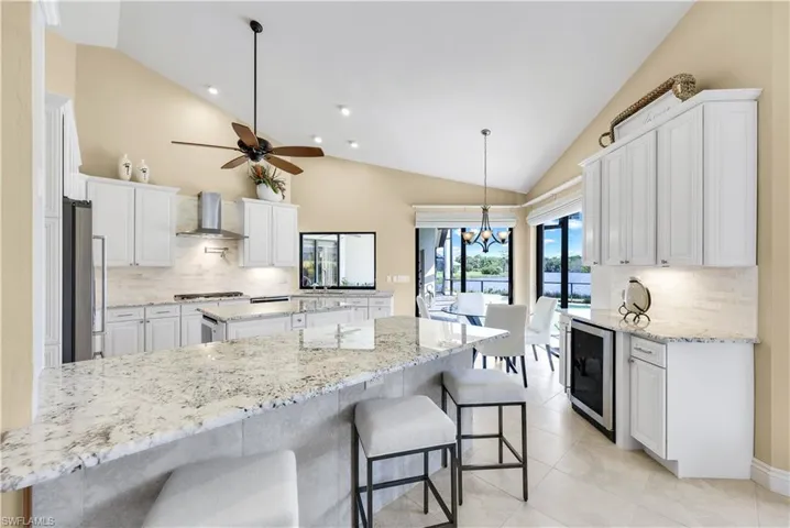 Kitchen with wine cooler, ceiling fan with notable chandelier, vaulted ceiling, white cabinetry, and wall chimney exhaust hood