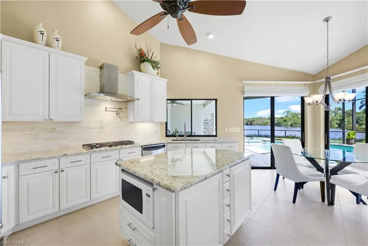 Kitchen featuring ceiling fan with notable chandelier, white cabinets, vaulted ceiling, wall chimney exhaust hood, and white microwave