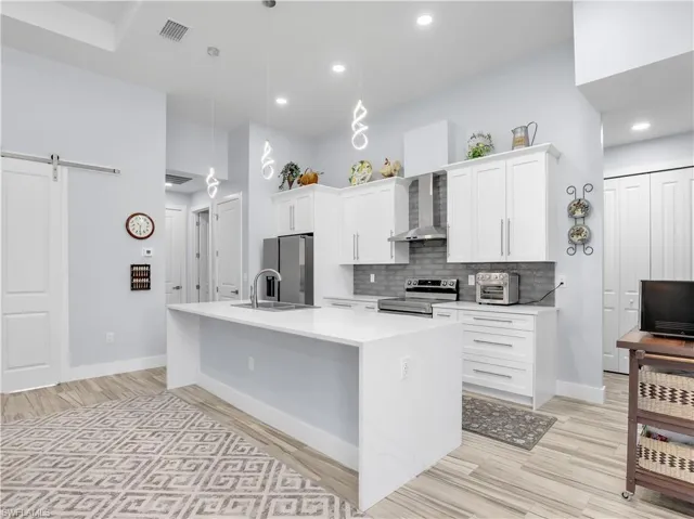 Kitchen featuring an island with sink, decorative light fixtures, wall chimney range hood, stainless steel appliances, and a barn door