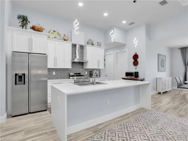 Kitchen with wall chimney exhaust hood, a kitchen island with sink, pendant lighting, appliances with stainless steel finishes, and white cabinetry