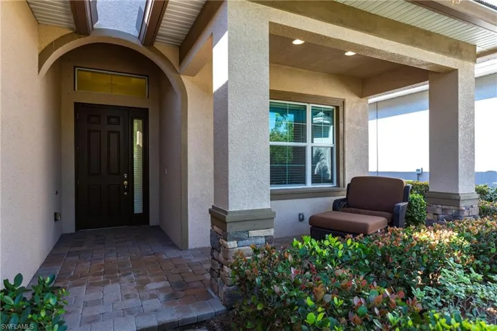 Inviting front entrance featuring a dark paneled door with a sidelight and transom window, a covered porch with recessed lighting, stone-clad pillars, and paver flooring
