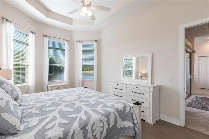 Bedroom featuring a tray ceiling with a ceiling fan, three windows with horizontal blinds and sheer curtains, wood-finish flooring, and a doorway opening to an adjacent hallway