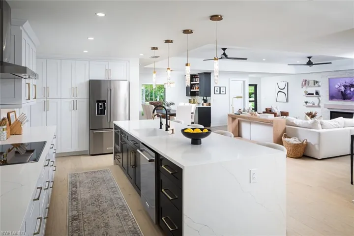 Kitchen featuring wall chimney exhaust hood, decorative light fixtures, stainless steel appliances, an island with sink, and white cabinetry