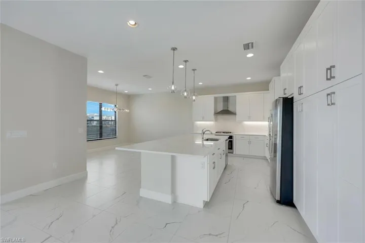 Kitchen with white cabinets, decorative light fixtures, wall chimney range hood, stainless steel appliances, and an island with sink