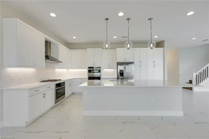 Kitchen with decorative light fixtures, white cabinetry, wall chimney range hood, stainless steel appliances, and a center island with sink
