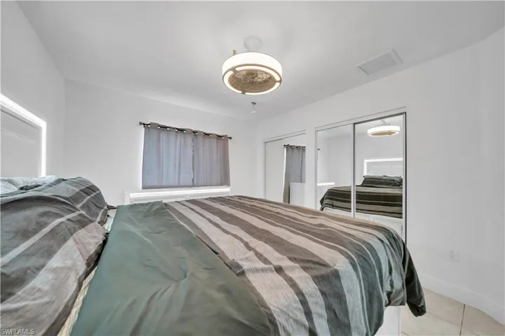 Crisp white walls frame this bedroom featuring a ceiling-mounted light fixture, sliding mirrored closet doors, a window with blinds, and tile flooring