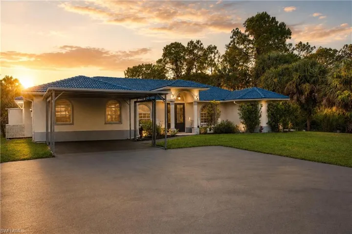 Front exterior showcasing the circular driveway, covered parking area, mature landscaping, and expansive Naples Florida homesite.