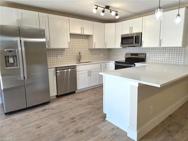 Modern kitchen featuring white shaker cabinetry, stainless steel appliances, a patterned tile backsplash, light-toned wood-finish flooring, and a spacious island with a light-colored countertop