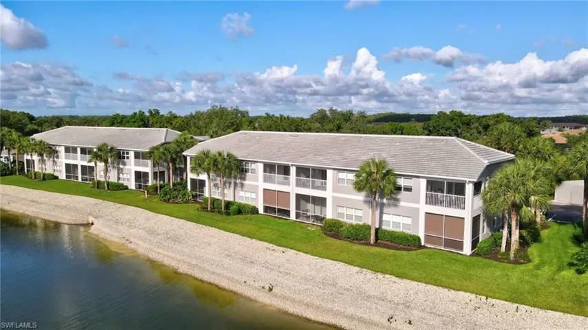 Waterfront multi-story buildings featuring screened patios and balconies, a waterside sandy bank, landscaped green space, and mature palm trees