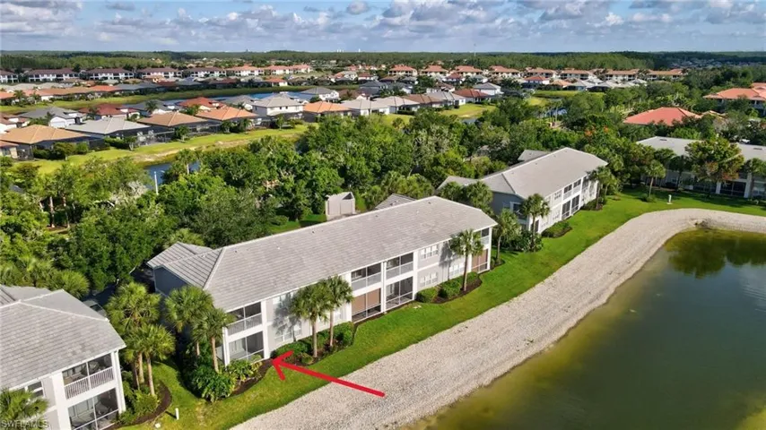 Waterfront condominium building featuring a tiled roof, screened balconies, mature palm trees, and manicured landscaping, situated beside a water feature and gravel bank