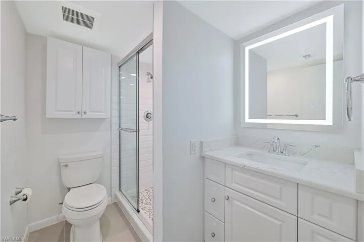 Contemporary bathroom featuring a spacious vanity with white cabinetry and a light-toned countertop, an illuminated mirror, and a glass-enclosed shower with subway tile surround and pebble-style flooring