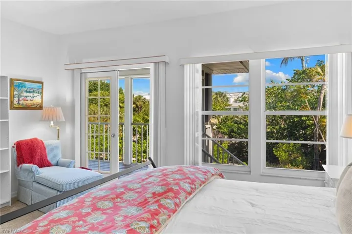 This inviting bedroom features light-colored walls, a window offering views of lush greenery and blue skies, and a sliding glass door opening to a private balcony