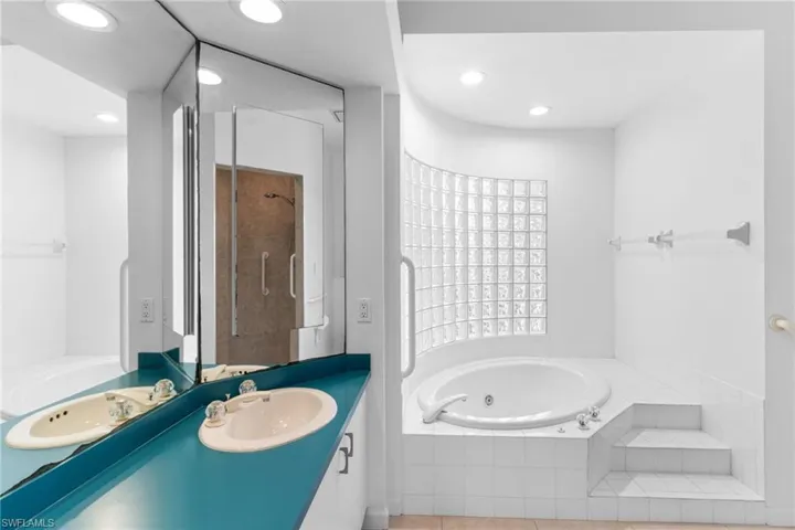 The bathroom includes a double vanity with a teal countertop and a spacious built-in tub with tile surround and steps, illuminated by recessed lighting