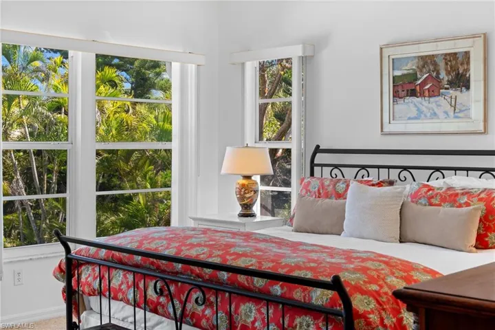 This bright room features white walls, a wrought iron bed frame, and large windows offering verdant outdoor views