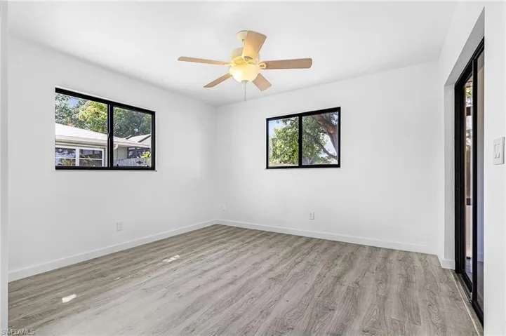 Spacious room featuring wood-finish flooring, a ceiling fan with integrated lighting, two black-framed windows, and a black-framed sliding glass door