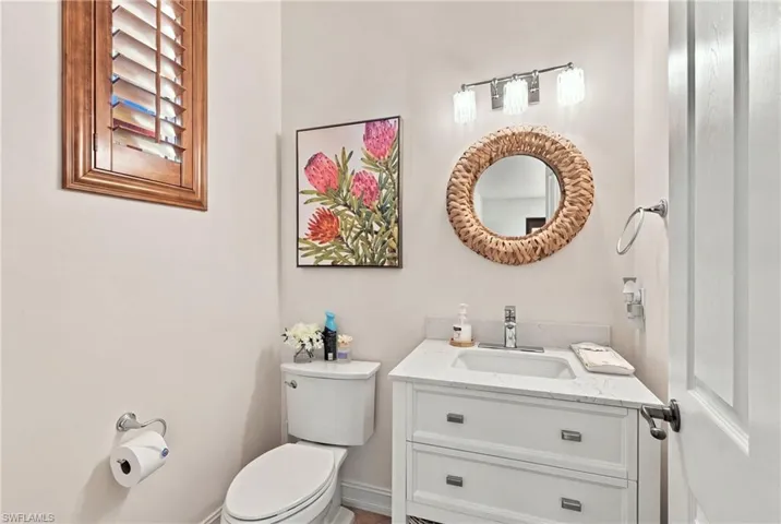 White vanity cabinet with a stone-top integrated sink, three-light brushed-nickel fixture, wood-framed louvered window shutter, and circular textured-frame mirror