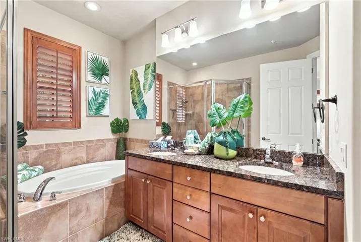 Dual-sink vanity with stone-finish countertop and wood cabinetry, soaking tub with tile surround, glass-enclosed shower, and plantation shutters