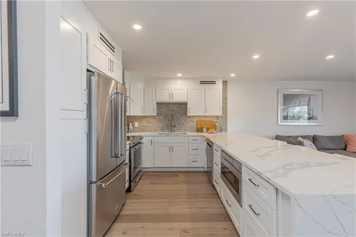 Modern kitchen featuring white shaker cabinetry, stainless steel appliances, a large island with a light-toned Quartz countertop, and light wood-finish flooring