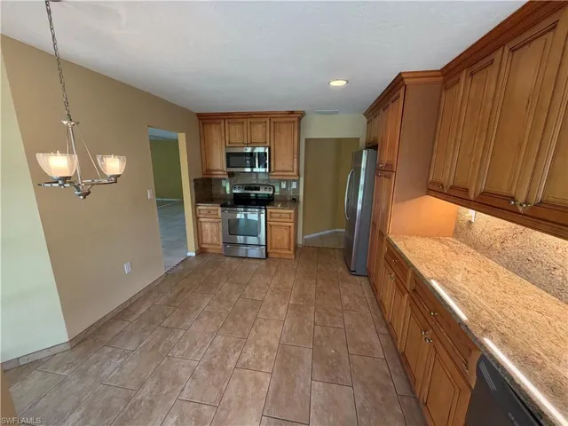 Kitchen with stainless steel appliances, wood finish cabinets, light stone countertops, hanging lights, and backsplash