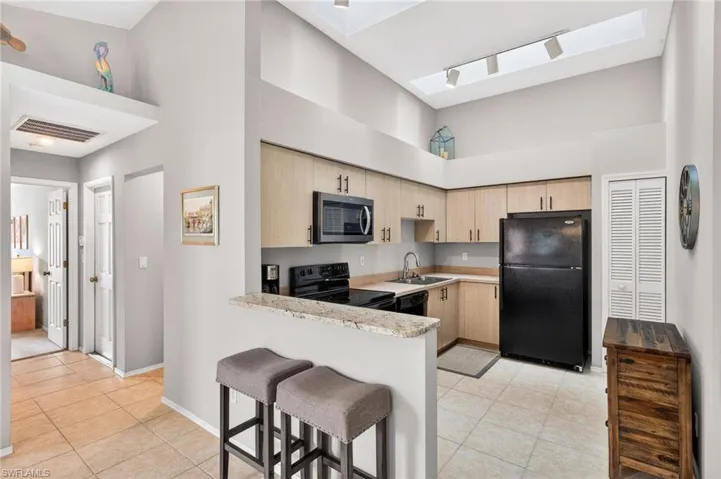 Kitchen featuring a high ceiling, black appliances, a skylight, light wood finish cabinetry, and a kitchen bar