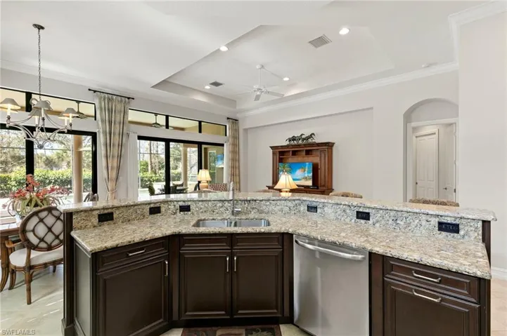Kitchen featuring dark wood finish cabinets, stainless steel dishwasher, ornamental molding, light stone countertops, and a tray ceiling