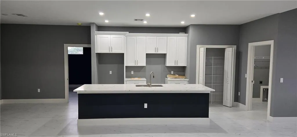 Kitchen with a center island with sink, light marble finish flooring, white cabinetry, a sink, and baseboards