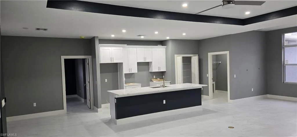 Kitchen with a kitchen island with sink, ceiling fan, white cabinetry, light marble finish flooring, and baseboards