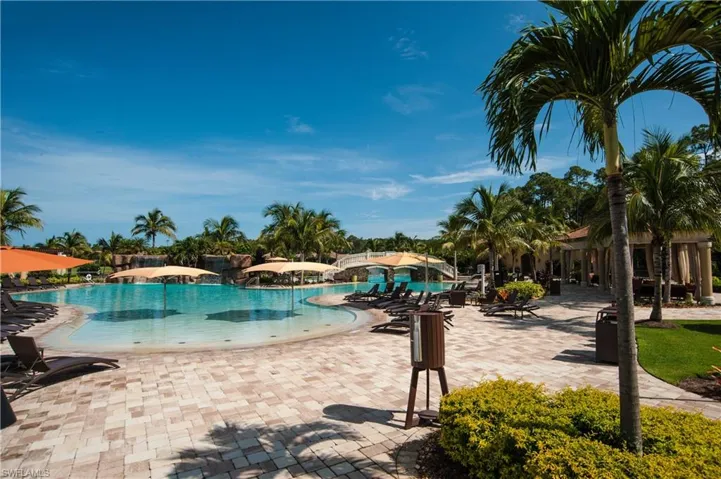 Expansive outdoor pool area featuring a large resort-style pool with a gradual entry, substantial paved patio, lush tropical landscaping with palm trees, and a decorative bridge