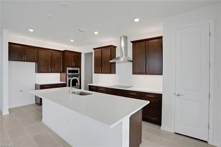 Kitchen featuring a kitchen island with sink, recessed lighting, stainless steel appliances, light tile patterned flooring, and dark wood finish cabinets