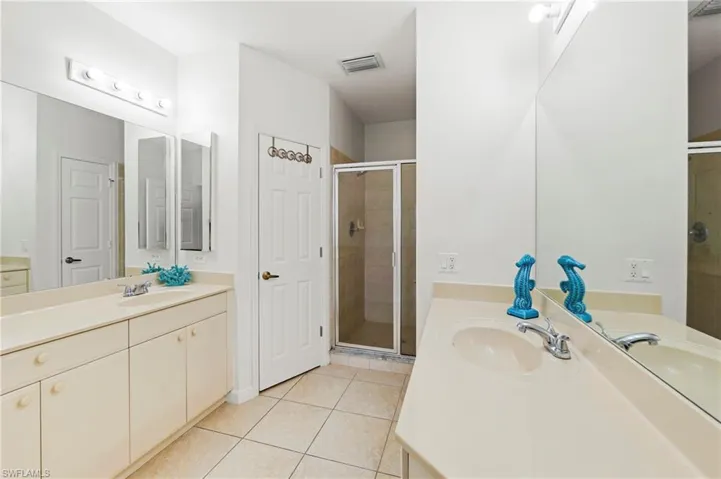 Bathroom featuring dual vanity sinks set into a light-toned countertop, a glass-enclosed shower, a built-in wall mirror, and neutral tile flooring