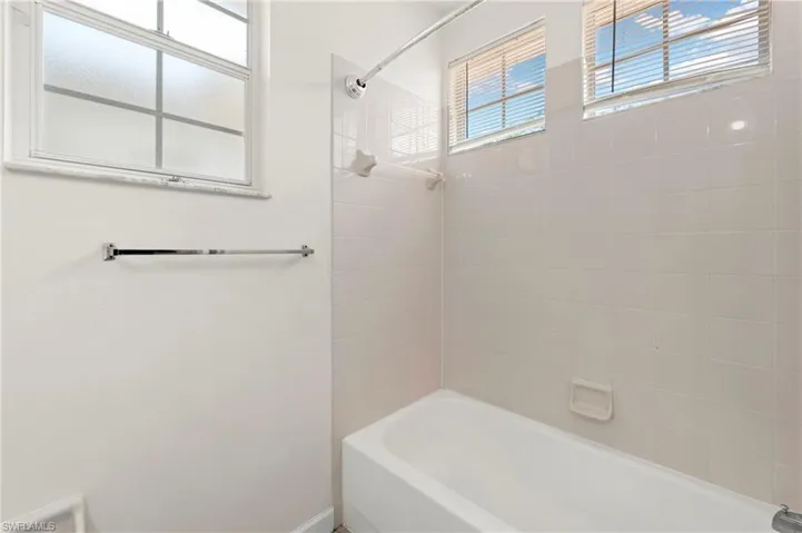 Bathroom with a white tub and shower combination, featuring white tile surround, two windows with horizontal blinds, and a frosted glass window