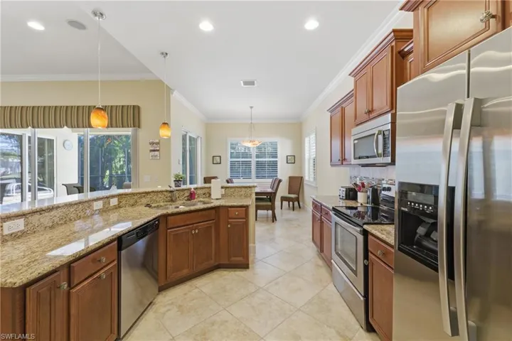 Kitchen featuring stainless steel appliances, light stone countertops, hanging light fixtures, ornamental molding, and brown cabinetry