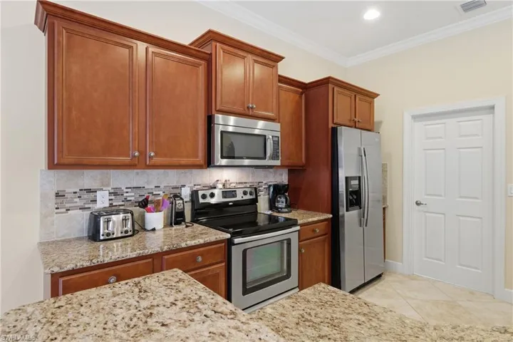 Kitchen with stainless steel appliances, brown cabinets, decorative backsplash, light stone counters, and ornamental molding
