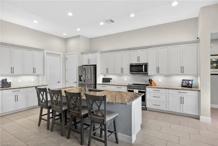 Kitchen with stainless steel appliances, a breakfast bar, light stone countertops, an island with sink, and white cabinets with under and upper cabinet lighting
